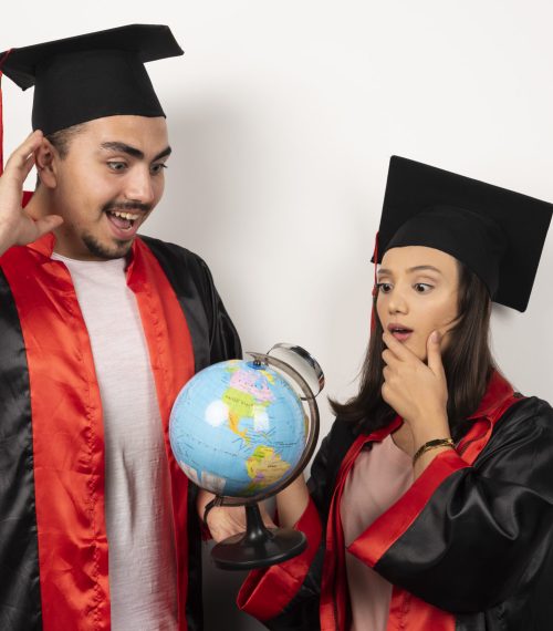 Pair of happy students in gown looking at globe on white background. High quality photo